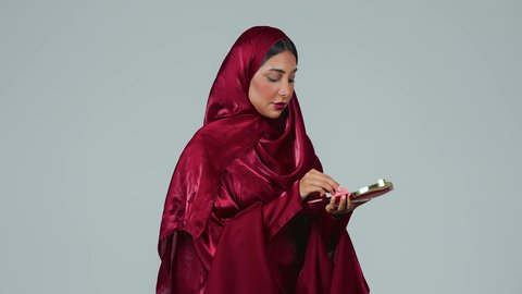 بورتريه لسيدة عربية خليجية سعودية سمراء ترتدي العباءة والحجاب تمسك بيديها علبة واسفنجة أحمر الخدود, النظر إلى الكاميرا بإيماءات السعادة والسرور, مفهوم التزين والتجمل, الاهتمام والعناية بالبشرة والجمال, الشغف بوضع المكياج,  خلفية رمادية