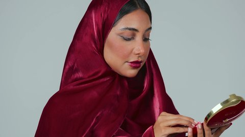 مفهوم التزين والتجمل, الاهتمام والعناية بالبشرة والجمال, الشغف بوضع المكياج,  بورتريه لسيدة عربية خليجية سعودية سمراء ترتدي العباءة والحجاب تمسك بيديها علبة واسفنجة أحمر الخدود, النظر إلى الكاميرا بإيماءات السعادة والسرور, خلفية رمادية