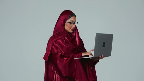 إنجاز المهام المطلوبة عبر اللابتوب، ادارة الأعمال عن بعد، استخدام الأجهزة الحديثة والمتطورة، بورتريه لامرأة عربية خليجية سعودية سمراء ترتدي العباءة والحجاب ونظارة طبية تمسك بيدها جهاز الحاسوب المحمول، دمج التكنولوجيا بالعمل،  مهن ووظائف نسائية، خلفية رمادية