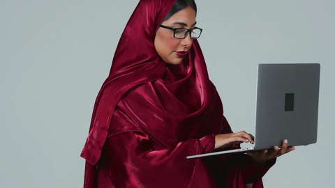Remote business management, using modern and advanced devices, a portrait of a Saudi Arabian Gulf woman with a dark complexion wearing an abaya and hijab and holding a laptop in her hand, integrating technology with work, completing required tasks via the laptop, women's professions and jobs, gray background.