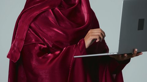 ادارة الأعمال عن بعد، استخدام الأجهزة الحديثة والمتطورة، بورتريه لامرأة عربية خليجية سعودية سمراء ترتدي العباءة والحجاب ونظارة طبية تمسك بيدها جهاز الحاسوب المحمول، دمج التكنولوجيا بالعمل، إنجاز المهام المطلوبة عبر اللابتوب، مهن ووظائف نسائية، خلفية رمادية