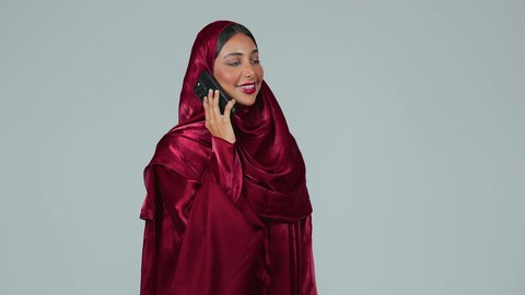 اجراء مكالمة هاتفية عبر الجوال، استخدام جهاز تقني حديث ومتطور، تصفح مواقع التواصل الاجتماعي، بورتريه لامرأة عربية خليجية سعودية سمراء ترتدي العباءة والحجاب تمسك بيدها الهاتف المحمول، خلفية رمادية