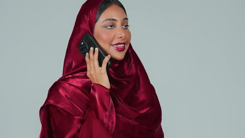 استخدام جهاز تقني حديث ومتطور، الدردشة مع الأصدقاء والأقارب عبر الجوال، تصفح مواقع التواصل الاجتماعي، بورتريه لامرأة عربية خليجية سعودية سمراء ترتدي العباءة والحجاب تمسك بيدها الهاتف المحمول، خلفية رمادية