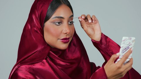 بورتريه لامرأة عربية خليجية سعودية سمراء ترتدي العباءة والحجاب تقوم بوضع الرموش الاصطناعية, مفهوم التزين والتجمل, التسويق لمنتجات ومستحضرات التجميل, الاناقة والاهتمام بالجمال والمظهر الخارجي, خلفية رمادية