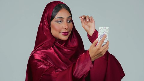 التسويق لمنتجات ومستحضرات التجميل, مفهوم التزين والتجمل,الاناقة والاهتمام بالجمال والمظهر الخارجي, بورتريه لامرأة عربية خليجية سعودية سمراء ترتدي العباءة والحجاب تقوم بوضع الرموش الاصطناعية, خلفية رمادية