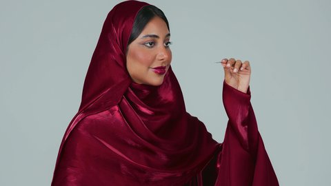 الاناقة والاهتمام بالجمال والمظهر الخارجي, مفهوم التزين والتجمل, التسويق لمنتجات ومستحضرات التجميل, بورتريه لامرأة عربية خليجية سعودية سمراء ترتدي العباءة والحجاب تقوم بوضع الرموش الاصطناعية, خلفية رمادية