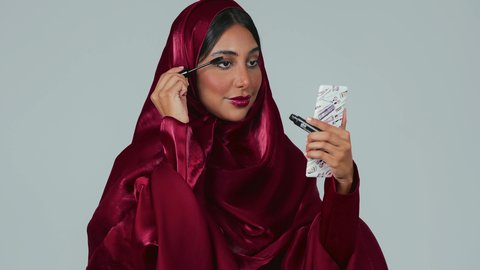 بورتريه لسيدة عربية خليجية سعودية سمراء ترتدي العباءة والحجاب تقوم بوضع الميكاب، استخدام الماسكرا لإبراز جمال العينين، الإهتمام بالجمال والمظهر الخارجي، دورات تعليمية لمكياج تتوريال, خبيرة مستحضرات التجميل النسائية، خلفيه رمادية