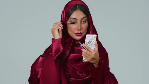 Using mascara to highlight the beauty of the eyes, caring about beauty and appearance, educational courses for makeup tutorials, a female cosmetics expert, a portrait of a Saudi Arabian Gulf woman with a dark complexion wearing an abaya and hijab applying makeup, gray background.