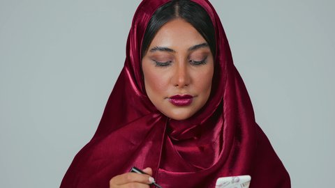 دورات تعليمية لمكياج تتوريال, استخدام الماسكرا لإبراز جمال العينين، الإهتمام بالجمال والمظهر الخارجي، خبيرة مستحضرات التجميل النسائية، بورتريه لسيدة عربية خليجية سعودية سمراء ترتدي العباءة والحجاب تقوم بوضع الميكاب، خلفيه رمادية