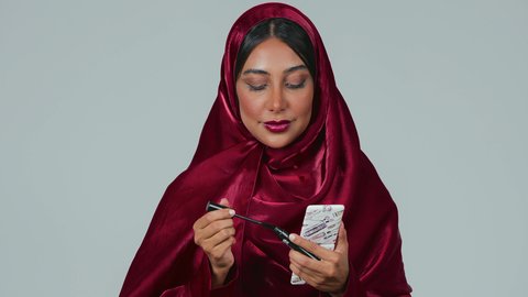 استخدام الماسكرا لإبراز جمال العينين، الإهتمام بالجمال والمظهر الخارجي، دورات تعليمية لمكياج تتوريال, خبيرة مستحضرات التجميل النسائية، بورتريه لسيدة عربية خليجية سعودية سمراء ترتدي العباءة والحجاب تقوم بوضع الميكاب، خلفيه رمادية