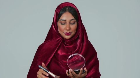 الشغف بوضع المكياج,  مفهوم التزين والتجمل, الاهتمام والعناية بالبشرة والجمال, بورتريه لسيدة عربية خليجية سعودية سمراء ترتدي العباءة والحجاب تمسك بيديها علبة وفرشاة أحمر الخدود, النظر إلى الكاميرا بإيماءات السعادة والسرور, خلفية رمادية