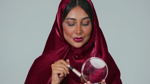 مفهوم التزين والتجمل, الاهتمام والعناية بالبشرة والجمال, الشغف بوضع المكياج,  بورتريه لسيدة عربية خليجية سعودية سمراء ترتدي العباءة والحجاب تمسك بيديها علبة وفرشاة أحمر الخدود, النظر إلى الكاميرا بإيماءات السعادة والسرور, خلفية رمادية