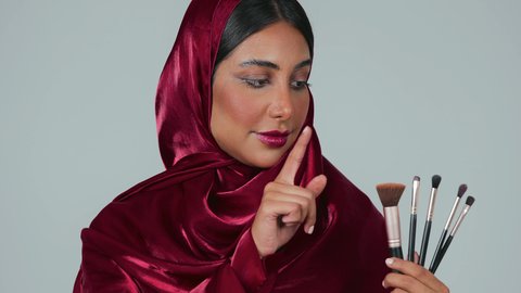 الاهتمام والعناية بالمظهر الخارجي, إيماءات النعومة والثقة، التسويق لأدوات ومنتجات التجميل, مفهوم التزين والتجمل, بورتريه لامرأة عربية خليجية سعودية سمراء ترتدي العباءة والحجاب تمسك فرش المكياج ، خلفية رمادية