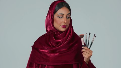 إيماءات النعومة والثقة، الاهتمام والعناية بالمظهر الخارجي, التسويق لأدوات ومنتجات التجميل, مفهوم التزين والتجمل, بورتريه لامرأة عربية خليجية سعودية سمراء ترتدي العباءة والحجاب تمسك فرش المكياج ، خلفية رمادية