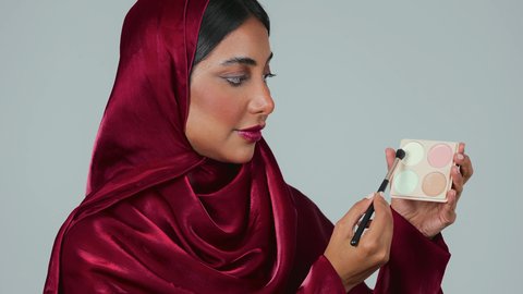 التسويق لمنتجات التجميل، أدوات مكياج تتوريال ، الإهتمام بالجمال والمظهر الخارجي ، بورتريه لامراة عربية خليجية سعودية سمراء ترتدي العباءة والحجاب تستخدم  فرشاة ظل العيون ، خلفية رمادية