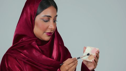 أدوات مكياج تتوريال ، الإهتمام بالجمال والمظهر الخارجي ، التسويق لمنتجات التجميل، بورتريه لامراة عربية خليجية سعودية سمراء ترتدي العباءة والحجاب تستخدم  فرشاة ظل العيون ، خلفية رمادية