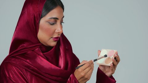 الإهتمام بالجمال والمظهر الخارجي ، أدوات مكياج تتوريال ،التسويق لمنتجات التجميل، بورتريه لامراة عربية خليجية سعودية سمراء ترتدي العباءة والحجاب تستخدم  فرشاة ظل العيون ، خلفية رمادية