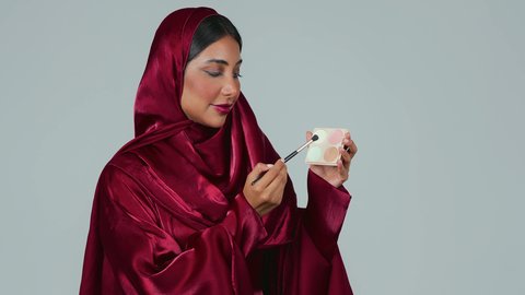 التسويق لمنتجات التجميل، أدوات مكياج تتوريال ، الإهتمام بالجمال والمظهر الخارجي ،بورتريه لامراة عربية خليجية سعودية سمراء ترتدي العباءة والحجاب تستخدم  فرشاة ظل العيون ، خلفية رمادية