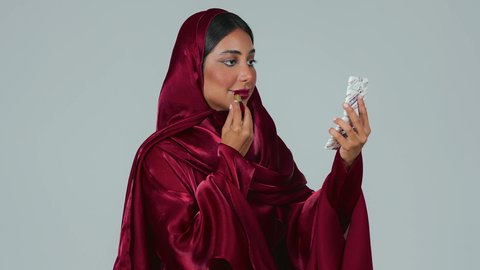 مفهوم الانوثة والنعومة ،  الإهتمام بالجمال والبشرة، ادوات مكياج تتوريال، بورتريه لمرأة عربية خليجية سعودية سمراء ترتدي العباءة والحجاب تحمل إحدى أدوات التجميل (قلم أحمر الشفاة) ، خلفية رمادية