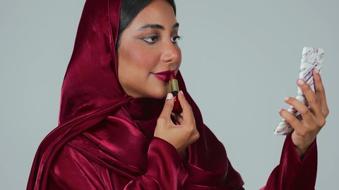 بورتريه لمرأة عربية خليجية سعودية سمراء ترتدي العباءة والحجاب تحمل إحدى أدوات التجميل (قلم أحمر الشفاة) ، مفهوم الإهتمام بالجمال والبشرة، ادوات مكياج تتوريال،خلفية رمادية