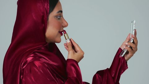 ادوات مكياج تتوريال، مفهوم الإهتمام بالجمال والبشرة،بورتريه لمرأة عربية خليجية سعودية سمراء ترتدي العباءة والحجاب تحمل إحدى أدوات التجميل (قلم أحمر الشفاة) ، خلفية رمادية