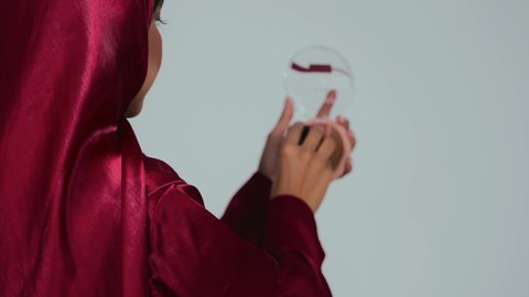 النظر  بإيماءات السعادة والسرور, مفهوم التزين والتجمل, الاهتمام والعناية بالبشرة والجمال, الشغف بوضع المكياج,  بورتريه لسيدة عربية خليجية سعودية سمراء ترتدي العباءة والحجاب تمسك بيديها علبة وفرشاة أحمر الخدود, خلفية رمادية