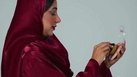 بورتريه لسيدة عربية خليجية سعودية سمراء ترتدي العباءة والحجاب تمسك بيديها علبة وفرشاة أحمر الخدود, النظر إلى الكاميرا بإيماءات السعادة والسرور, مفهوم التزين والتجمل, الاهتمام والعناية بالبشرة والجمال, الشغف بوضع المكياج, خلفية رمادية