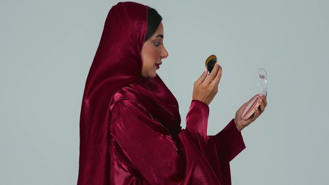 الشغف بوضع المكياج, مفهوم التزين والتجمل, الاهتمام والعناية بالبشرة والجمال,  بورتريه لسيدة عربية خليجية سعودية سمراء ترتدي العباءة والحجاب تمسك بيديها علبة وفرشاة أحمر الخدود, النظر إلى الكاميرا بإيماءات السعادة والسرور, خلفية رمادية