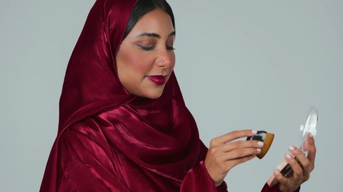 مفهوم التزين والتجمل, الاهتمام والعناية بالبشرة والجمال, الشغف بوضع المكياج,  بورتريه لسيدة عربية خليجية سعودية سمراء ترتدي العباءة والحجاب تمسك بيديها علبة وفرشاة أحمر الخدود, النظر إلى الكاميرا بإيماءات السعادة والسرور, خلفية رمادية