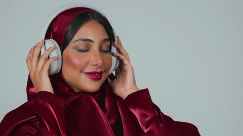 الاهتمام بالأناقة والمظهر الخارجي ، الاستماع الى الأغاني والموسيقى عبر سماعة الرأس، تعابير وايماءات السرور والسعادة، بورتريه لسيدة عربية خليجية سعودية سمراء ترتدي العباءة والحجاب تضع الهيدفون، خلفية رمادية
