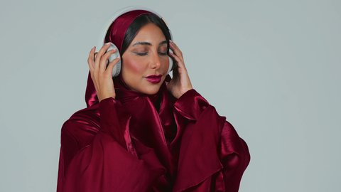 A portrait of a dark-skinned Saudi Arabian Gulf woman wearing an abaya and hijab, with headphones on, showing an interest in elegance and appearance, listening to songs and music through the headphones, with expressions and gestures of joy and happiness, against a gray background.