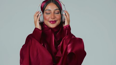 تعابير وايماءات السرور والسعادة، الاستماع الى الأغاني والموسيقى عبر سماعة الرأس،بورتريه لسيدة عربية خليجية سعودية سمراء ترتدي العباءة والحجاب تضع الهيدفون، الاهتمام بالأناقة والمظهر الخارجي ، خلفية رمادية