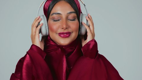 الاستماع الى الأغاني والموسيقى عبر سماعة الرأس، تعابير وايماءات السرور والسعادة، بورتريه لسيدة عربية خليجية سعودية سمراء ترتدي العباءة والحجاب تضع الهيدفون، الاهتمام بالأناقة والمظهر الخارجي ، خلفية رمادية