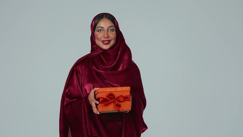 تقديم الهدايا بالاعياد والمناسبات الخاصة، مفهوم الشكر والامتنان، التعبير عن الحب والتقدير والاحترام،  بورتريه لسيدة عربية خليجية سعودية سمراء ترتدي العباءة والحجاب تحمل صندوق هدية احمر اللون  بايماءات السعادة والسرور، خلفية رماية