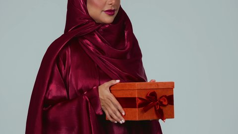 مفهوم الشكر والامتنان، تقديم الهدايا بالاعياد والمناسبات الخاصة، التعبير عن الحب والتقدير والاحترام،  بورتريه لسيدة عربية خليجية سعودية سمراء ترتدي العباءة والحجاب تحمل صندوق هدية احمر اللون  بايماءات السعادة والسرور، خلفية رماية