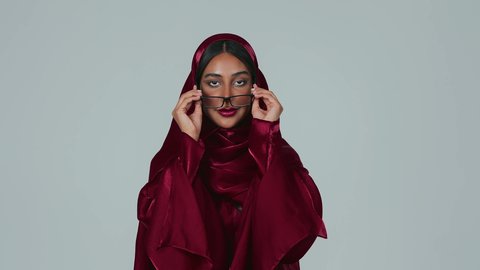 مفهوم تحسين وتعديل الرؤية، المحافظة على صحة العينين، ارتداء النظارات الخاصة بضعف النظر، بورتريه لسيدة عربية خليجية سعودية سمراء ترتدي العباءة والحجاب تقوم بوضع النظاره الطبيه ، خلفية رمادية