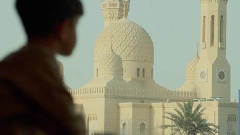 أماكن لأداء العبادة والتقرب إلى الله تعالى، تأمل معالم الوطن، الفن الهندسي المعماري للمساجد في الإمارات، تصوير مقرب من الخلف لولد عربي خليجي اماراتي يرتدي الكندورة يقوم بالنظر إلى المسجد