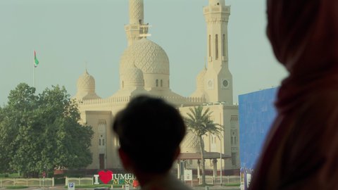 The architectural art of mosques in the UAE, places for worship and getting closer to Allah Almighty, reflecting the landmarks of the homeland, a close-up shot from behind of an Emirati Gulf Arab boy wearing a kandura standing with his mother as they look at the mosque.