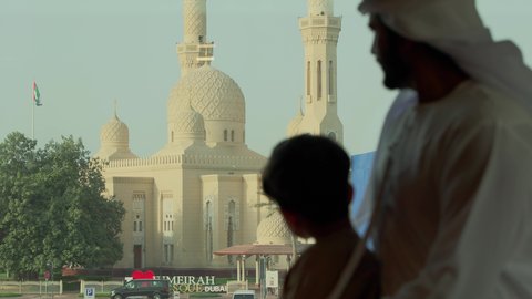 سارية علم الامارات ترفرف في سماء الوطن، الفن الهندسي المعماري للمساجد في الإمارات، أماكن لأداء العبادة والتقرب إلى الله تعالى، تصوير مقرب من الخلف لرجل عربي خليجي اماراتي يرتدي الكندورة والغترة يقف مع ابنه ويقوم بالنظر إلى المسجد