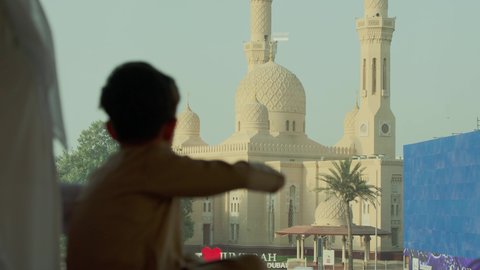 أماكن لأداء العبادة والتقرب إلى الله تعالى، الاناقة والاهتمام بالمظهر الخارجي، تصوير مقرب من الجانب لرجل عربي خليجي اماراتي يرتدي الكندورة والغترة الاماراتية ييقف مع ابنه ويقوم بالنظر إلى المسجد، الفن الهندسي المعماري للمساجد في الإمارات