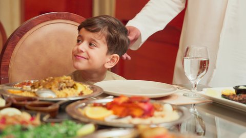 قضاء الأوقات الممتعة مع الابناء, تقديم الاهتمام والعناية للاولاد، مائدة مليئة بالاطباق والمأكولات الشعبية الشهية، أنشطة ترفيهية مسلية, الاستمتاع بالأجواء العائلية في الخارج, ولد عربي خليجي اماراتي يرتدي المندورة الاماراتية يجلس على طاولة الطعام في إحدى المطاعم