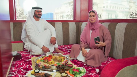 The love between spouses, restaurants for Eastern cuisine, culinary arts and recipes of Emirati cuisine, a mansaf of white rice with roasted chicken and spices garnished with vegetables in one of the Emirati restaurants, an Emirati Arab man wearing the kandura and ghutrah is enjoying delicious traditional dishes with his wife.