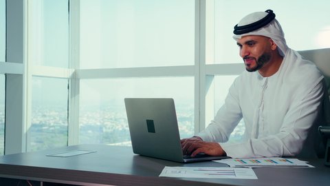 The development of Emirati companies, office tasks in companies in the United Arab Emirates, a close-up shot of an Emirati Arab man wearing a kandura and a white ghutrah using a laptop, accomplishing remote work tasks, managing and organizing business affairs via the laptop, with a background of towers and skyscrapers.