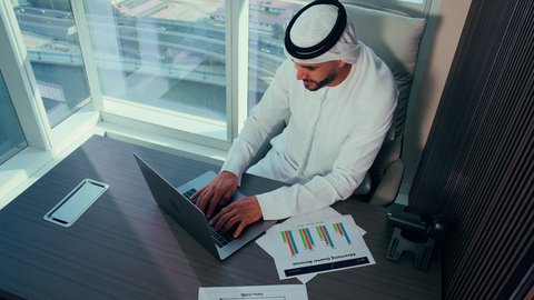 إتمام مهام العمل في المكتب،  مهام مكتبية في شركات الامارات العربية المتحدة، تصوير مقرب لرجل عربي خليجي اماراتي يرتدي الكندورة والغترة البيضاء يستخدم الحاسوب المحمول، إنجاز مهام العمل عن بعد، إدارة شؤون الأعمال وتنظيمها عبر اللابتوب، خلفية ابراج وناطحات سحاب