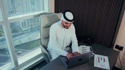 مهام مكتبية في شركات الامارات العربية المتحدة، تصوير مقرب لرجل عربي خليجي اماراتي يرتدي الكندورة والغترة البيضاء يستخدم الحاسوب المحمول، إنجاز مهام العمل عن بعد، إدارة شؤون الأعمال وتنظيمها عبر اللابتوب، ايماءات السعادة والسرور، خلفية ابراج وناطحات سحاب