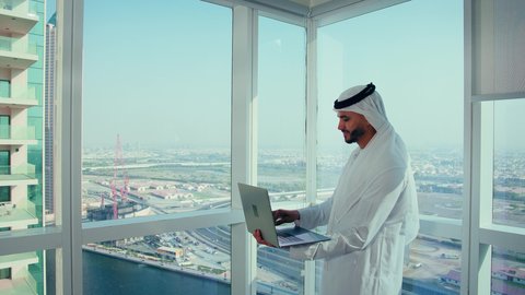 Office job in companies in the United Arab Emirates, close-up shot of an Arab Gulf Emirati man wearing a kandura and a white ghutrah using a laptop, completing remote work tasks, managing and organizing business affairs via the laptop, gestures of happiness and joy, background of towers and skyscrapers.