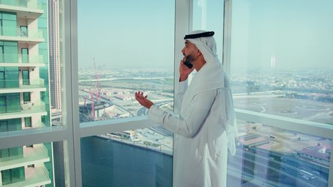 سماع الأخبار الجيدة عن المشاريع، شركات الامارات العربية المتحدة، تصوير مقرب لايدي رجل عربي خليجي اماراتي يرتدي الكندورة والغترة البيضاء يحمل هاتف محمول، إنجاز مهام العمل عن بعد، إدارة شؤون الأعمال وتنظيمها عبر اللابتوب، خلفية ابراج وناطحات سحاب