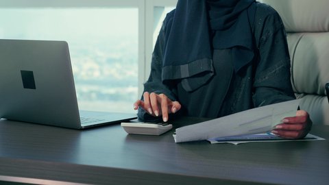 Using the calculator at work, auditing and analyzing project data, monitoring the chart plan, a close-up shot of an Emirati Gulf Arab woman analyzing data on a laptop, economic growth and financial investment, a Saudi woman wearing a hijab and abaya, background of skyscrapers.