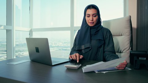 الآلة الحاسبة لانجاز مهام العمل، تدقيق وتحليل بيانات المشروع، متابعة مخطط الرسم البياني، امراه عربيه خليجيه اماراتيه تقوم بتحليل البيانات على الحاسوب المحمول، النمو الاقتصادي والاستثمار المالي، سيدة سعودية ترتدي الحجاب والعباءة، خلفية ناطحات سحاب