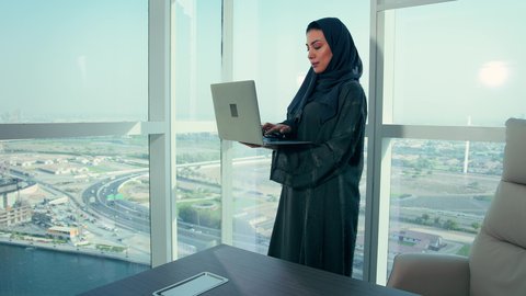 Studying new projects, auditing and analyzing project data, following the chart plan, an Emirati Gulf Arab woman analyzing data on a laptop, economic growth and financial investment, a Saudi woman wearing a hijab and abaya, office professions and jobs, background of towers and skyscrapers.